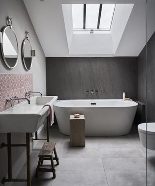 A bathroom with a double sink, grey floor and wall tiles and a large bath