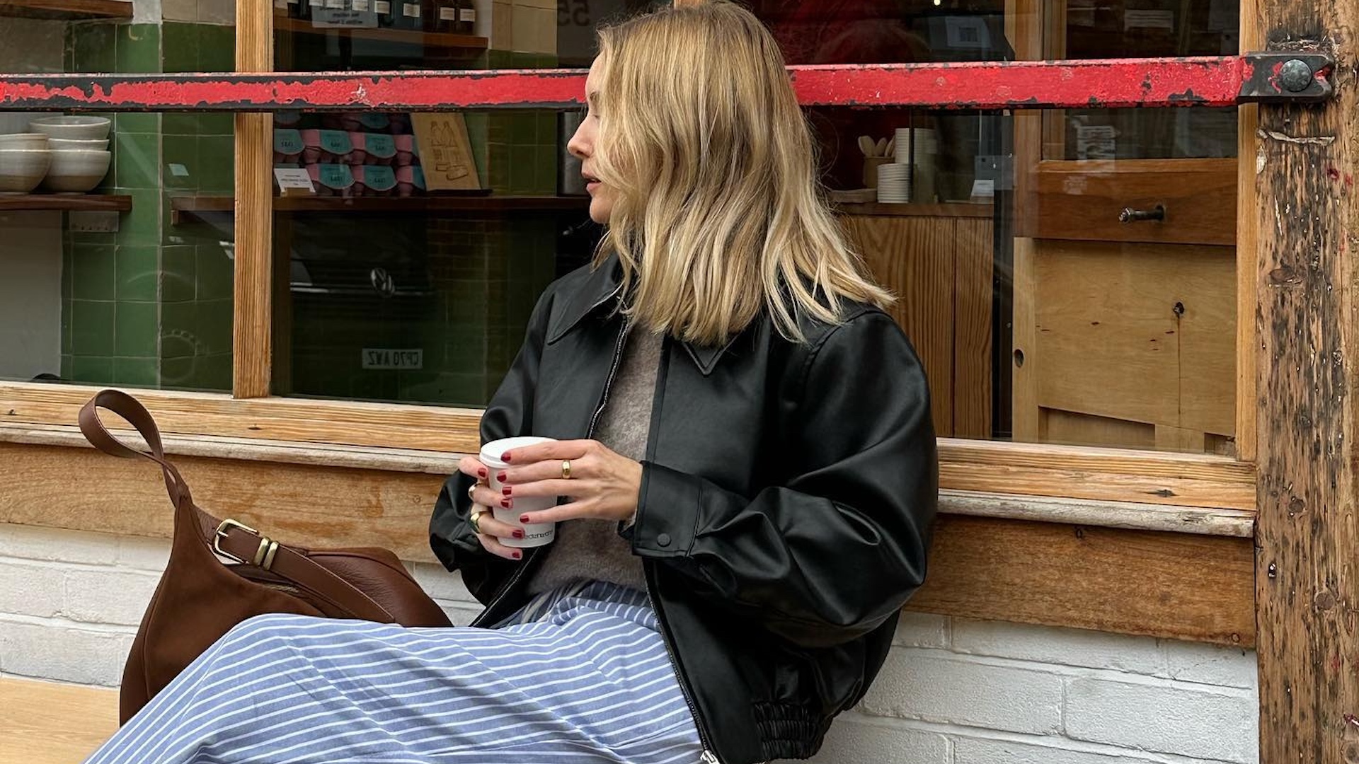 British influencer Chloe Butler sitting on a bench outside a London cafe holding a coffee wearing an oversize black leather jacket, neutral sweater, striped blue pajama pants, and a tan leather bag.