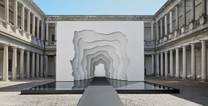 Daytime, outside image of the Divided Layers installation by Daniel Arsham, courtyard area, white stone building with stone columns around the edge, gravel floor, grey pathway, clear blue sky