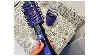 A person holding the purple Conair The Knot Dr. Blow Dryer brush with an included attachment sitting on the counter in the background, for the Conair blow dryer brush review.