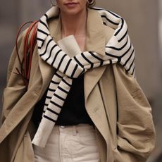 Dutch fashion influencer Anouk Yve poses wearing an oversize trench coat, a striped sweater over her shoulders, a black top, ivory jeans, and a strappy shoulder bag