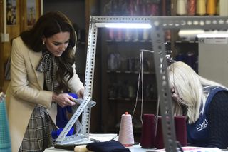 Kate Middleton wearing a white coat and black checked dress holding a pair of blue socks and leaning over a silver bar as a woman works on a knitting machine