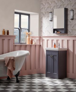 A bathroom with a windowsill with a peach vase with flowers, white walls, light pink wall panels, a navy blue sink unit with a mirrored medicine cabinet above it, a standing white bath, and gray and white flooring