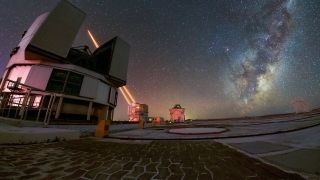 A foreground of curved bricks laid in a path leads to a round building with large section of roof opened. Behind it, another cylindrical building, shooting to brilliantly bright beams of light into the dark starry night sky. On the right, above the horizon, the arm of the Milky Way leans toward the top corner.