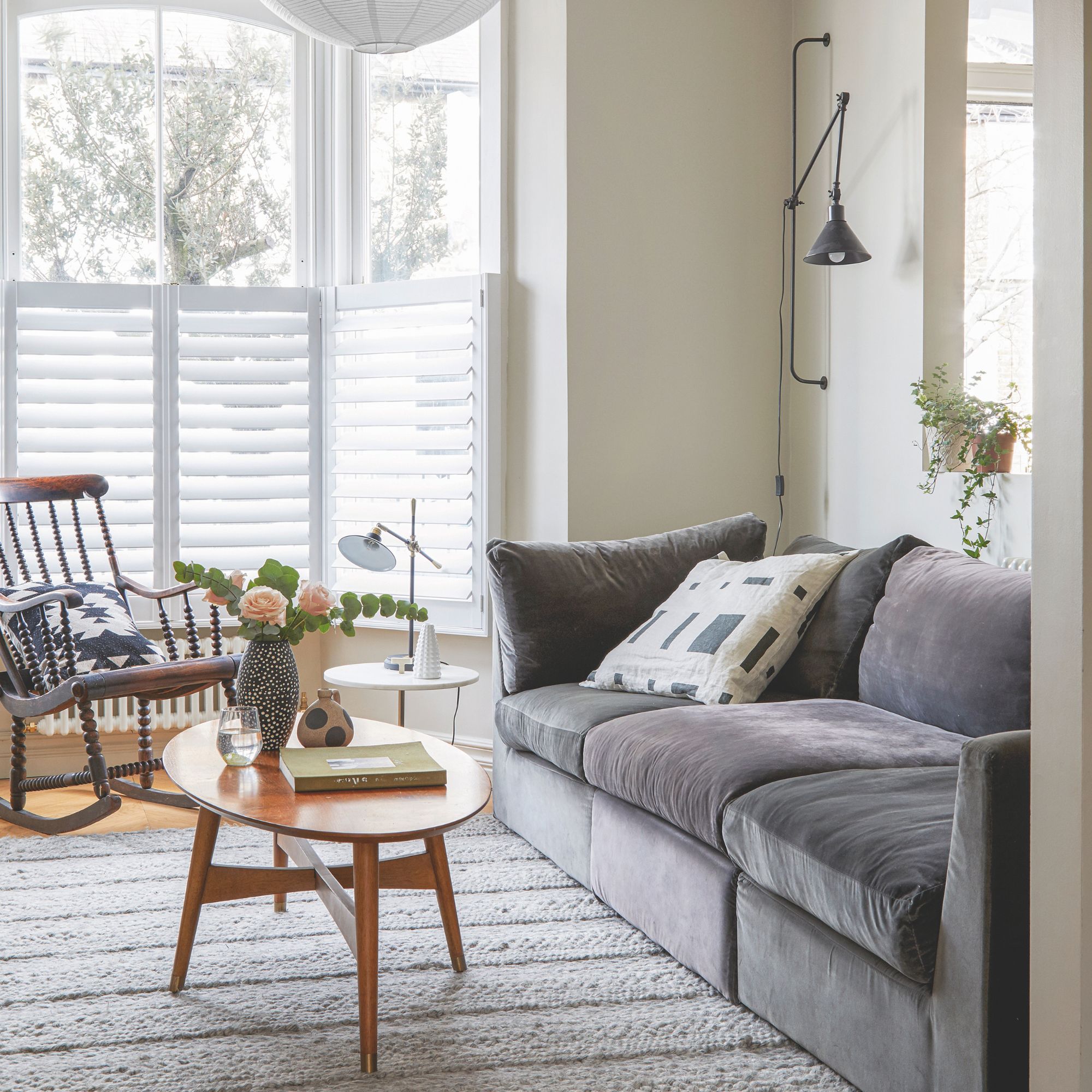 A white painted living room with a large bay window and plantation shutters. In front is a grey sofa with a wooden coffee table