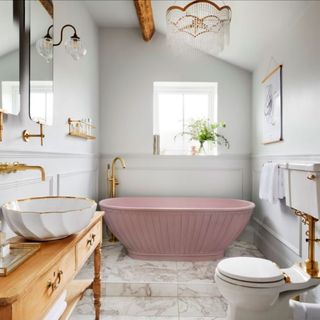 a bathroom with white walls and white marble floor tiles, aith a pink freestanding bathtub at the back, with gold chrome tap and pipe fittings