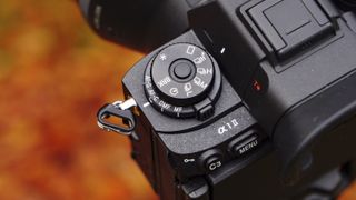 Closeup of the Sony A1 II mirrorless camera's drive mode dial, outdoors on a tripod with autumn leaves background