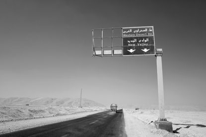 A single carriage way road with desert on either side an a road sign above with directions to Western Desert Rd and New Valley.