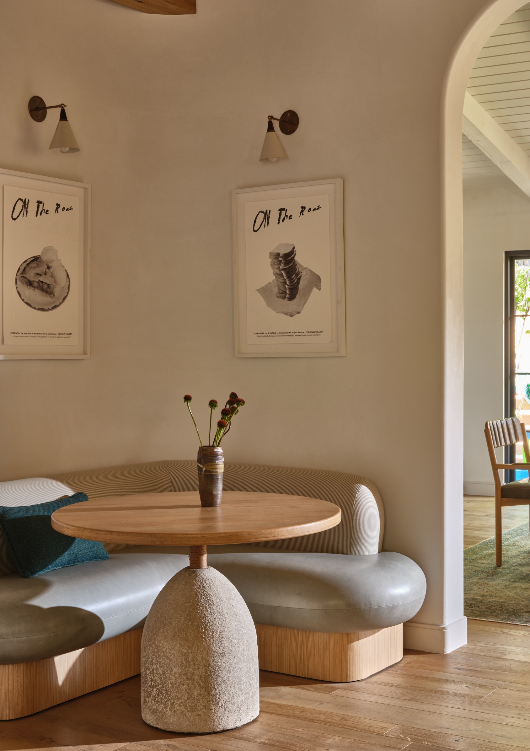 A modern banquette with large gray curved cushioned seating on a wooden base with a pedestal table with a stone base and round wooden top with two pieces of art on each corner above with a wall light