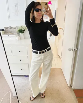 Judith wears white wide-leg jeans, a black belt, a black long-sleeve shirt, and black strappy heeled sandals.