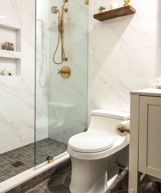 A white bathroom with a glass shower with a gold shower head and built in shelves, a white toilet with a dark wooden shelf with plants on over it, a white unit next to it, and gray marbled flooring