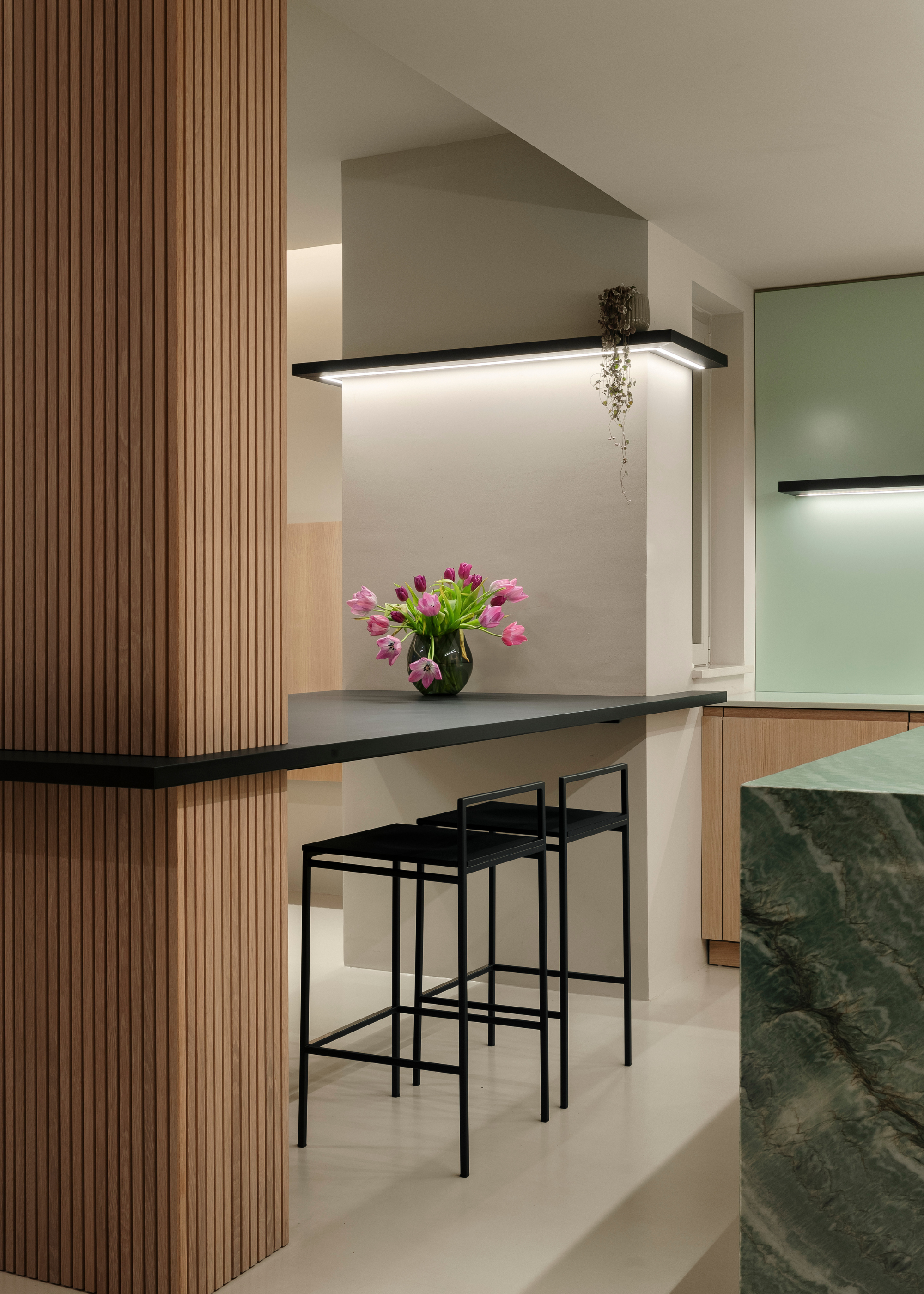 A kitchen with white walls, a vase of pink flowers by a pair of black kitchen stools by a green island