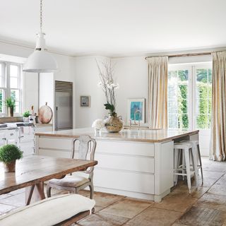 Large kitchen-diner with white kitchen island, a wooden dining room table, and cream curtains over the double doors to the garden