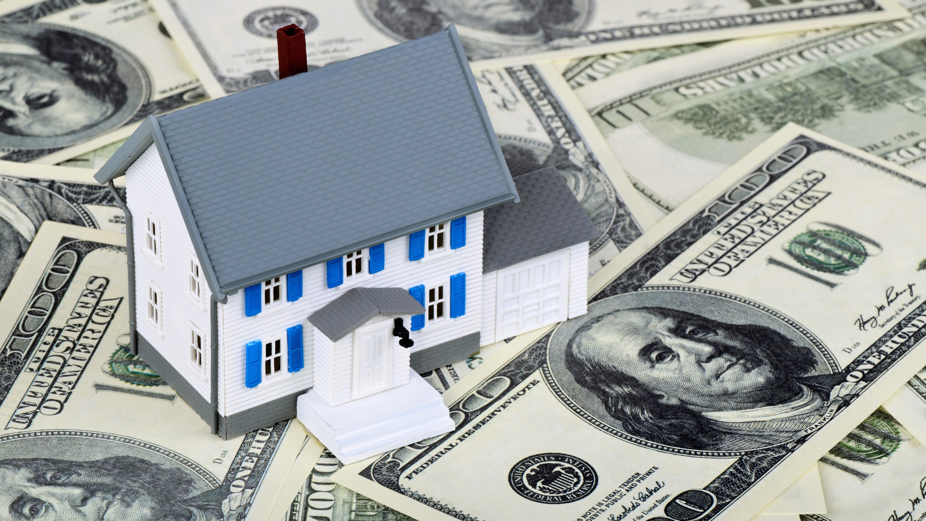 A toy home sits on top of scattered hundred-dollar bills.