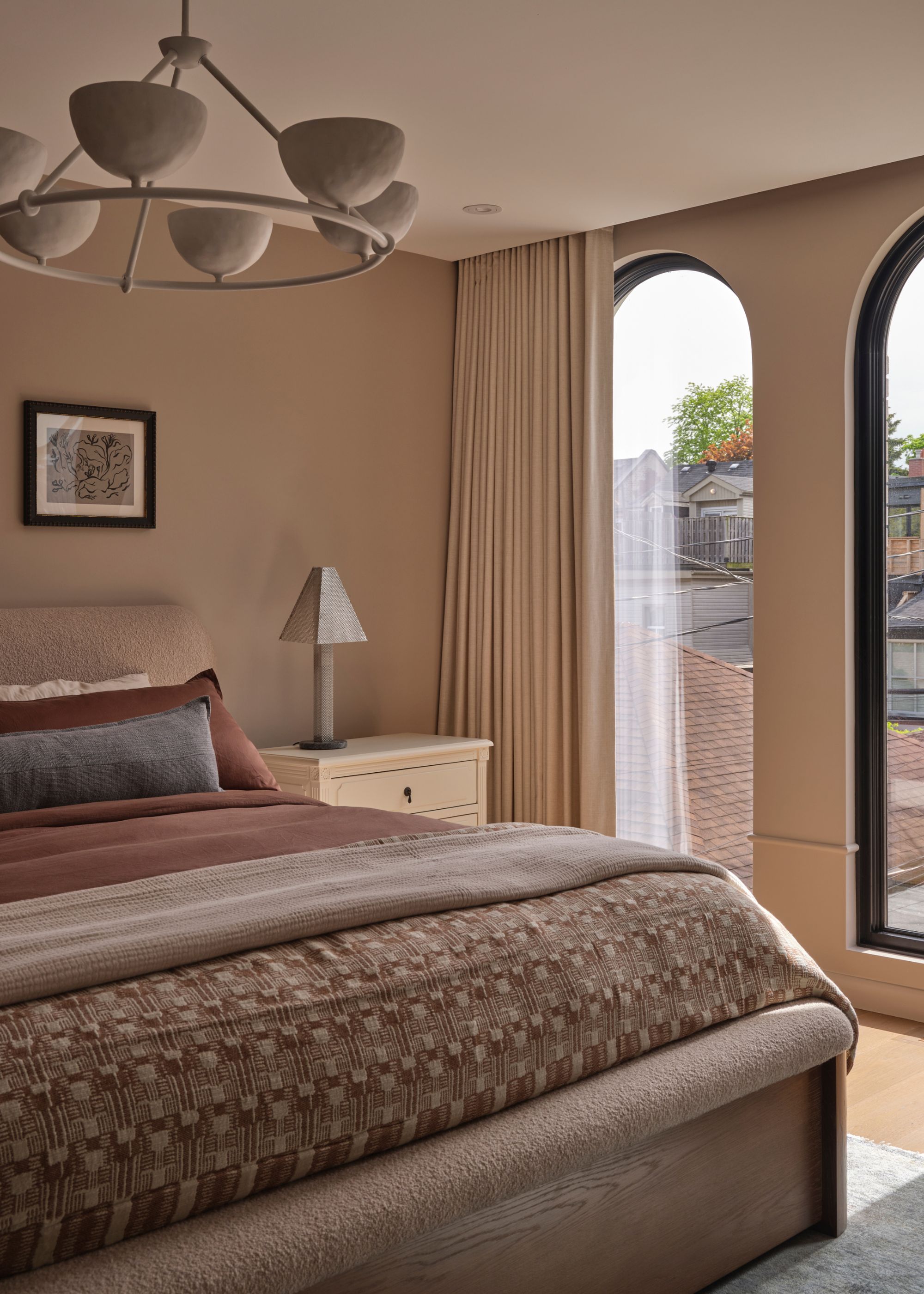 A bright bedroom with arched windows, a white nightstand, a statement lantern, and a layered bedscape with a patterned quilt