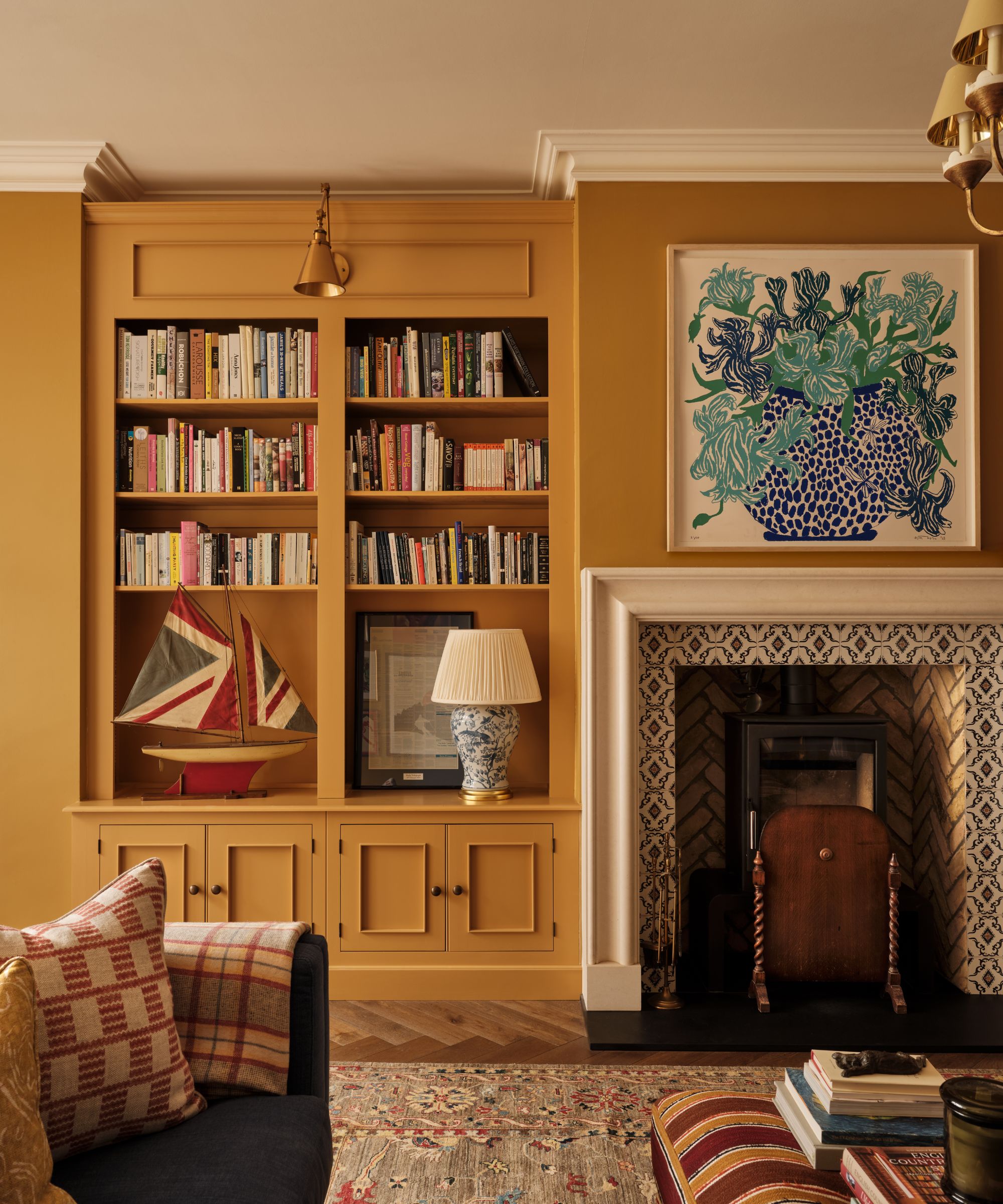 Living room with mustard yellow walls and painted bookshelves, a tiled fireplace with a log-burner, a blue still life painting of a vase of flowers above the fireplace and a striped ottoman