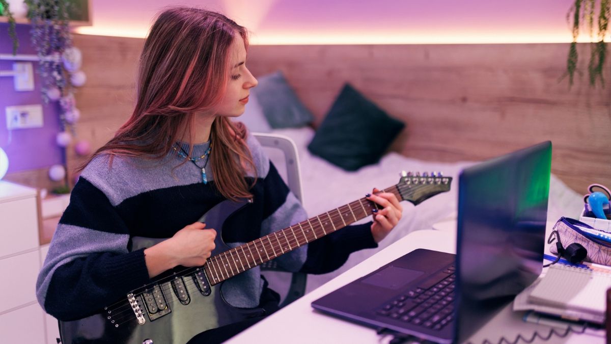 A woman plays electric guitar in front of a laptop