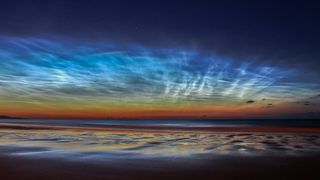 Wispy white noctilucent clouds swirl over an orange sunset and blue sea