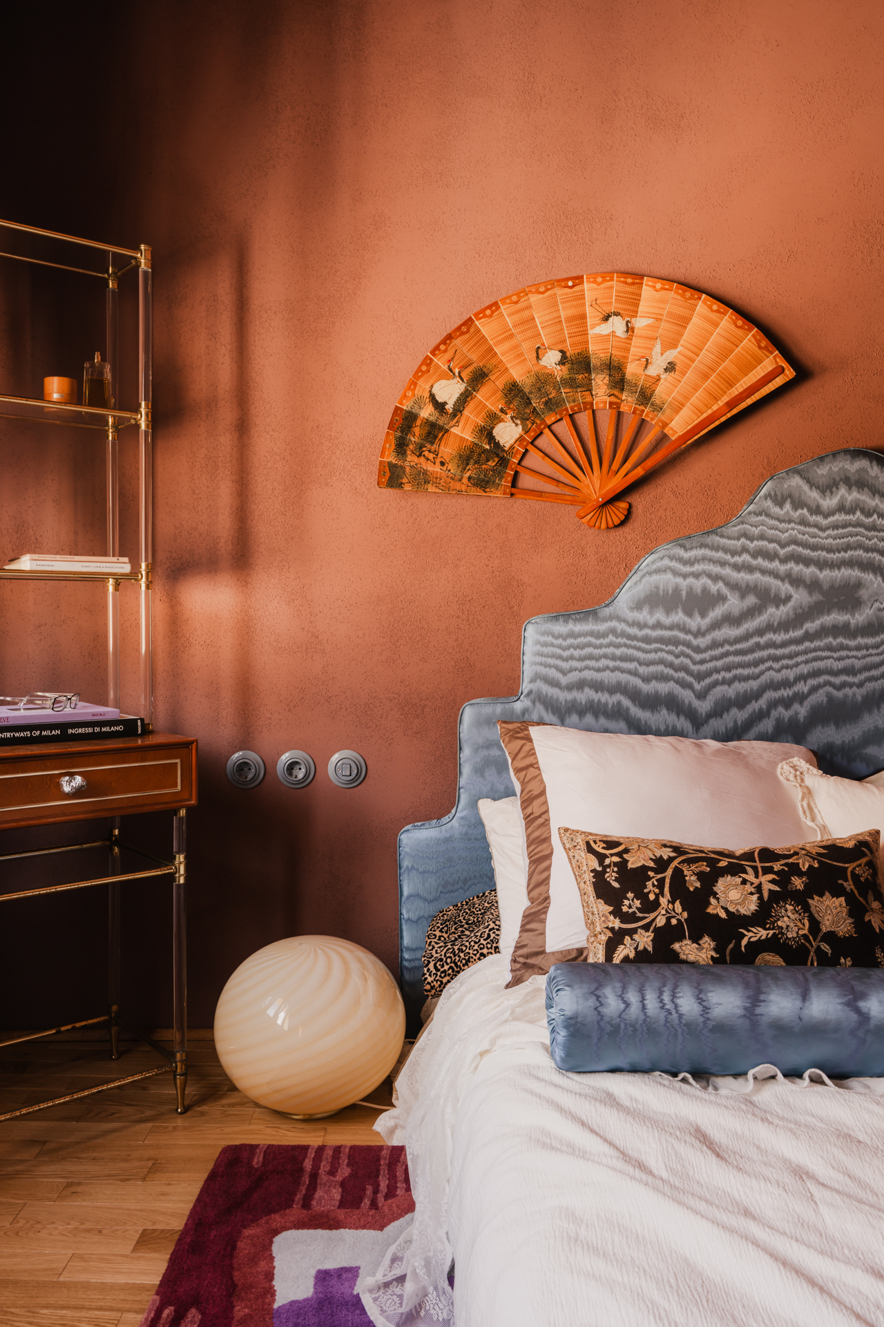 A blue headboard covered in moire jacquard, and a wooden fan hangs above the bed.
