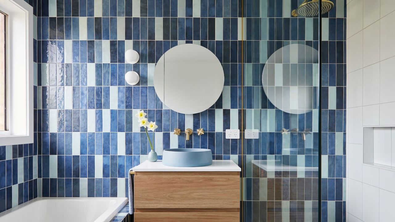 a tiled bathroom wth various shades of blue tiles, a blue basin and a wood vanity. a built in bath and shower enclosure are in view. 