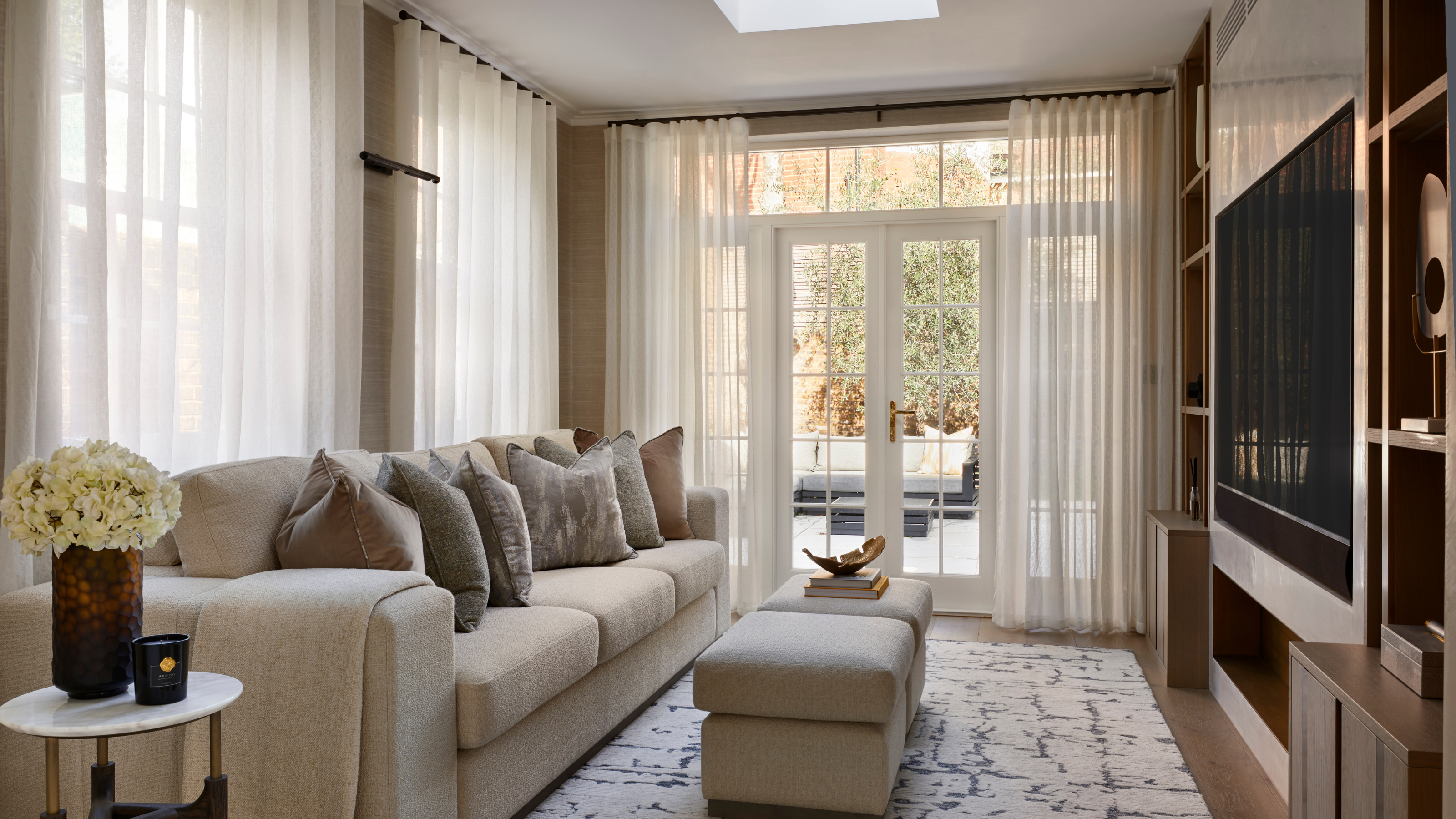 a living room with a light tan couch, several pillows, a pair of ottomans, and large windows covered by white sheer curtains