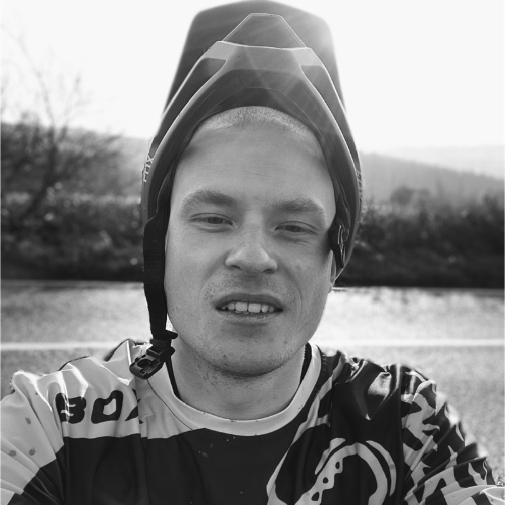 Black-and-white photo of a man wearing a mountain bike helmet pushed back and a patterned cycling jersey outdoors.