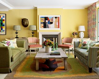 Colorful living room with yellow painted wall, two green sofas, two red chairs, low wooden coffee table, colorful artwork mounted above fireplace, three table lamps on console table