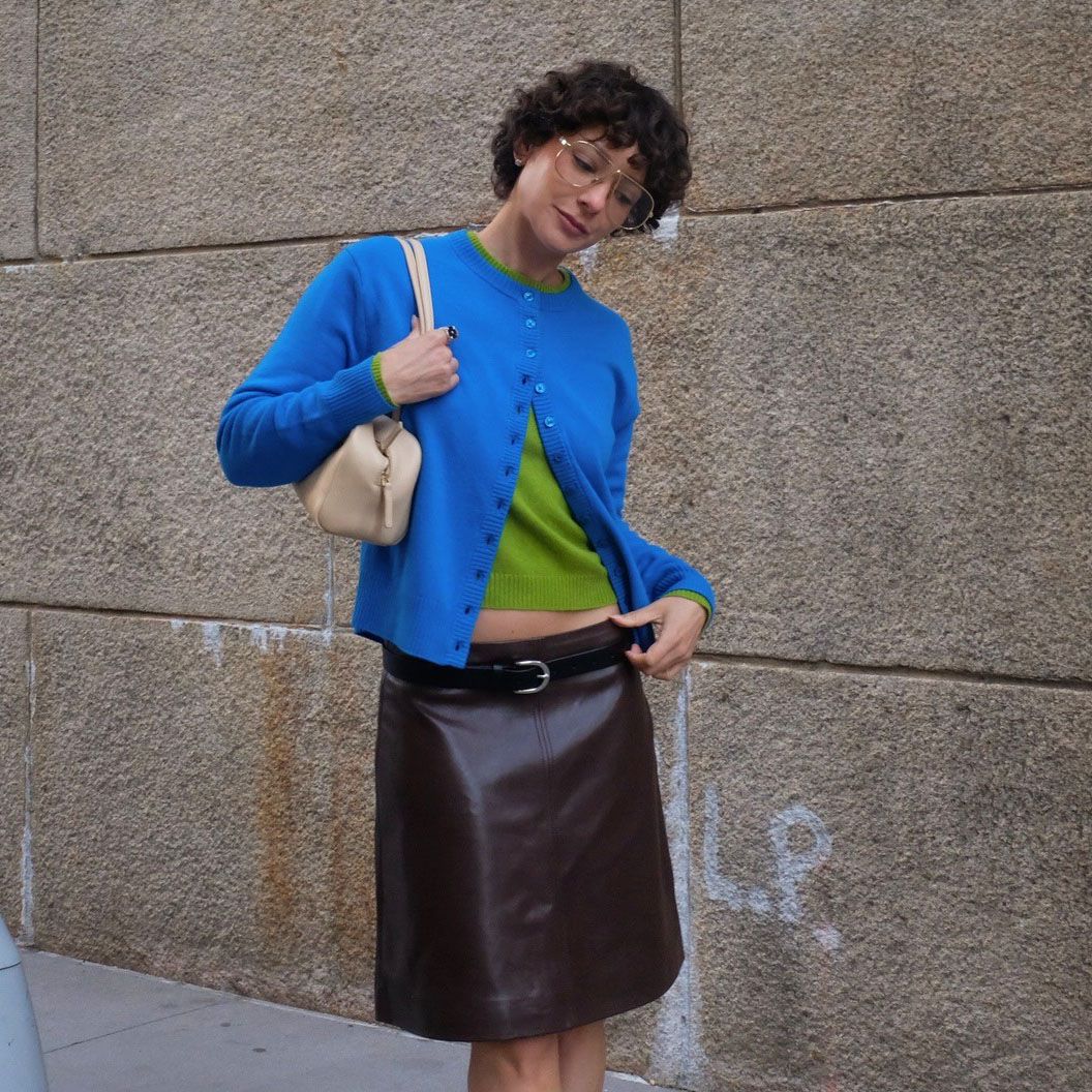 a woman wearing a blue cardigan, green t-shirt, brown leather skirt, and a tan shoulder bag