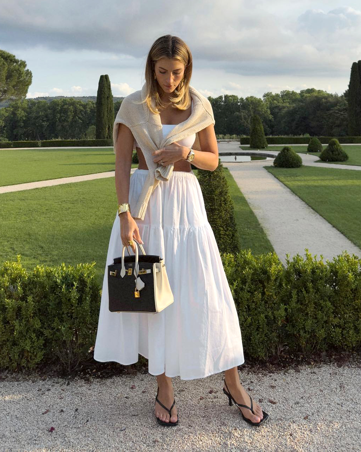 influencer Cass DiMicco poses in a garden in the south of France wearing a beige sweater over her shoulders, a white bandeau top, Hermes bag, full white tiered skirt, and slingback flip-flop kitten-heel sandals