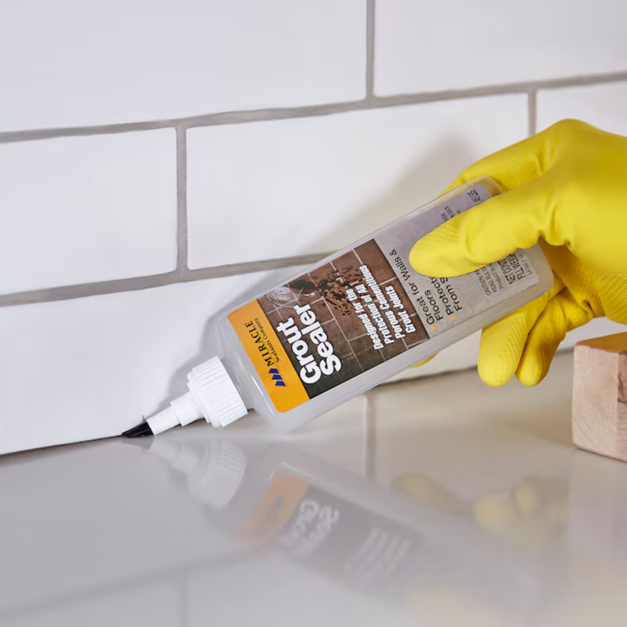 Bottle of grout sealer from Lowe's being used by a gloved hand to seal the gap between a floor and vertical tiled wall