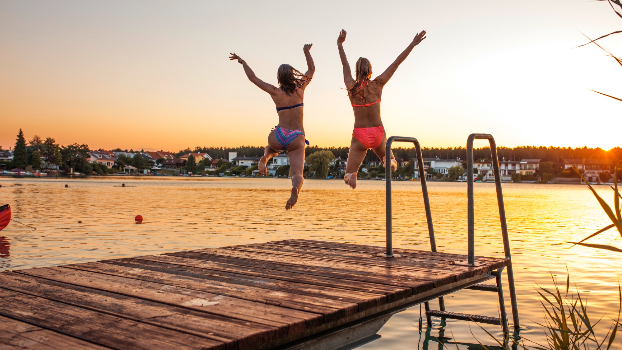 Two women wearing bathing suits jump into the water from a dock at sunset