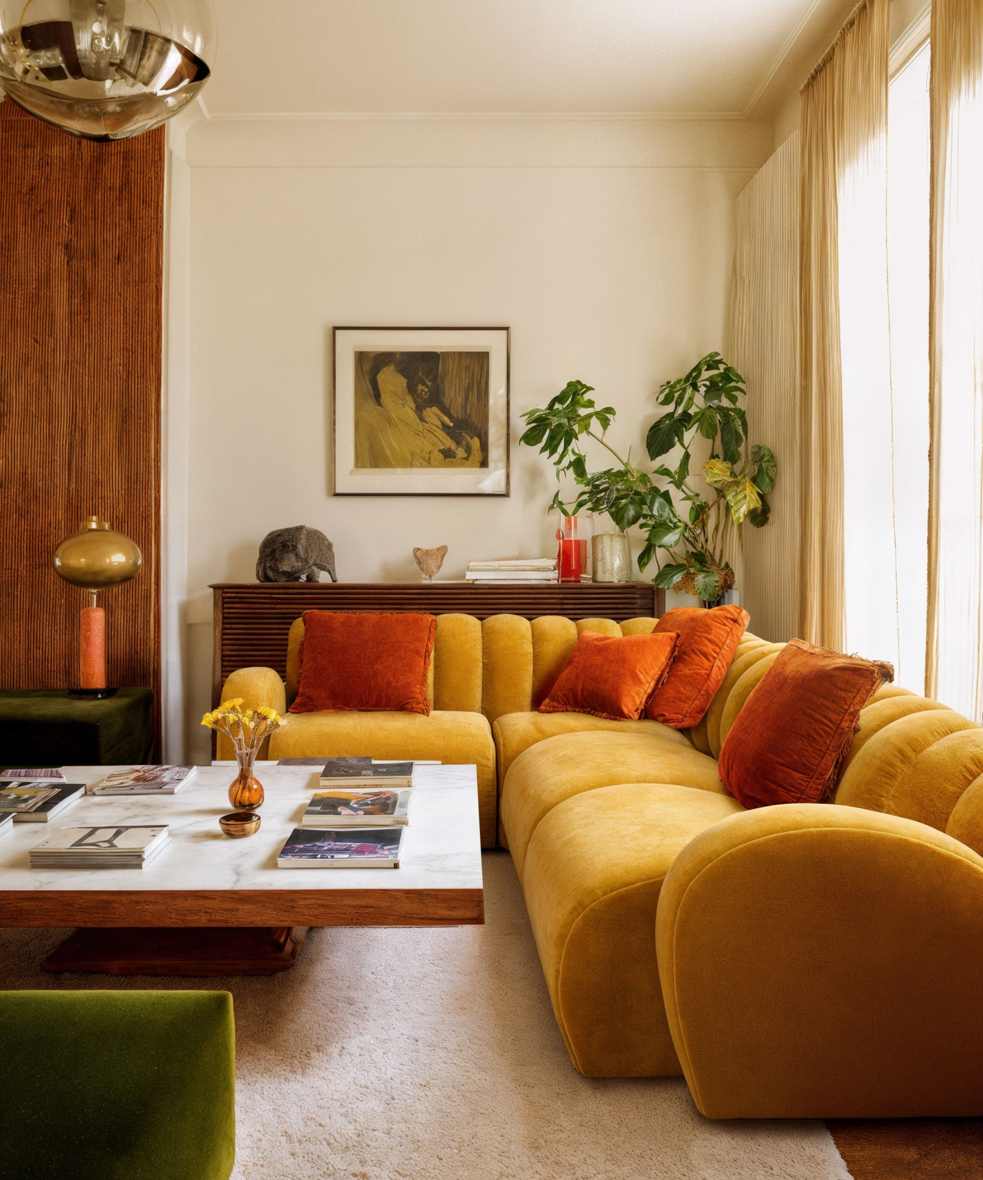 Mustard velvet sofa with orange cushions, framed art, plants and vintage sideboard in warm living room.