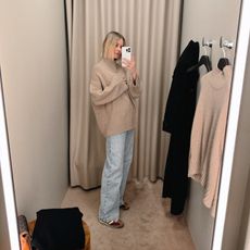 Natalie Cantell taking a dressing room selfie in a COS store in New York, wearing faded blue wide-leg jeans and an oversized beige sweater.