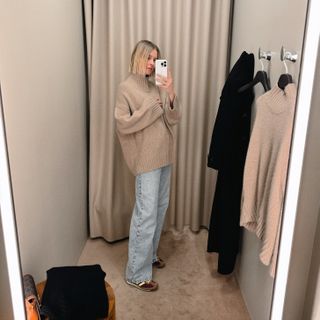 Natalie Cantell taking a dressing room selfie in a COS store in New York, wearing faded blue wide-leg jeans and an oversized beige sweater.