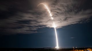 a rocket carves an arc of flame into a cloudy night sky during a launch.