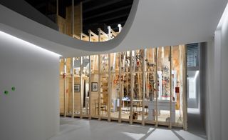 View of Forte’s room installation - a light wood structure offering a view of inside between the gaps. Chairs, a table and wall art can be seen inside along with an upper level. The pathway and walls around the installation are light grey