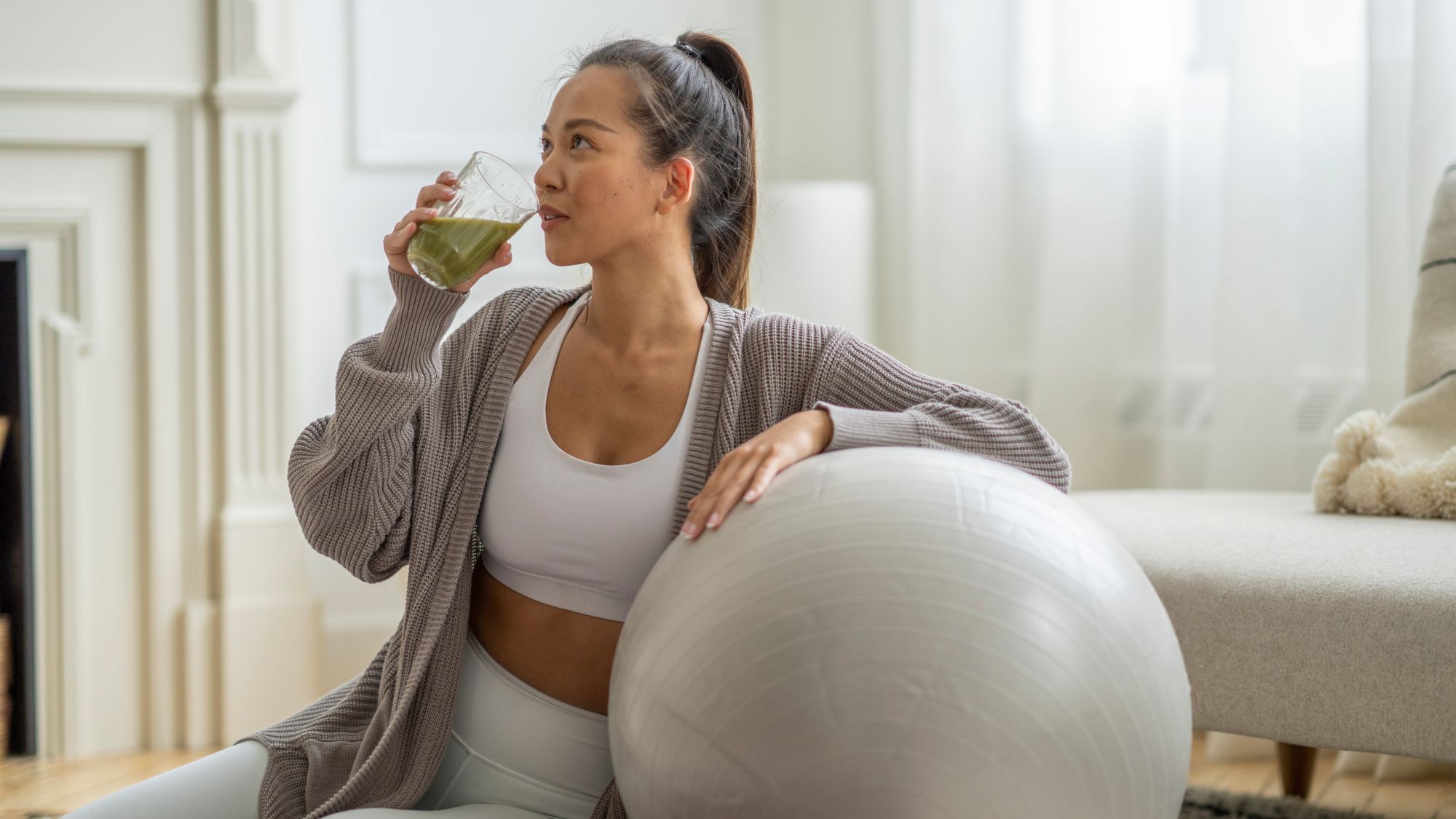 A woman drinking a smoothie in workout gear leaning on a workout ball
