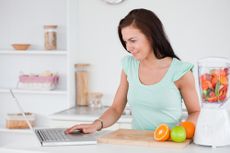 A woman prepares food in her kitchen and looks at her laptop.