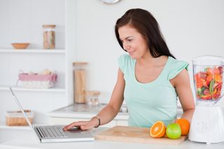 A woman prepares food in her kitchen and looks at her laptop.