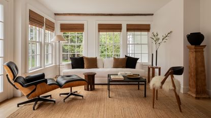 modern living room with bamboo blinds, a white sofa with caramel and black throw pillows, an eames lounge chair, a square coffee table and side table, an armchair with a sheepskin, a pedestal with a vase styled on top, and a jute rug on the floor