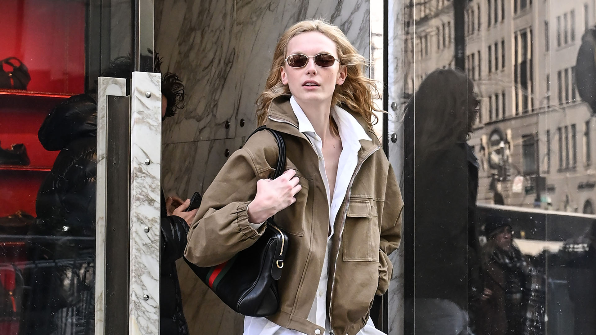 Alex Consani wearing a brown jacket, white button-down, blue jeans, black loafers, and black shoulder bag