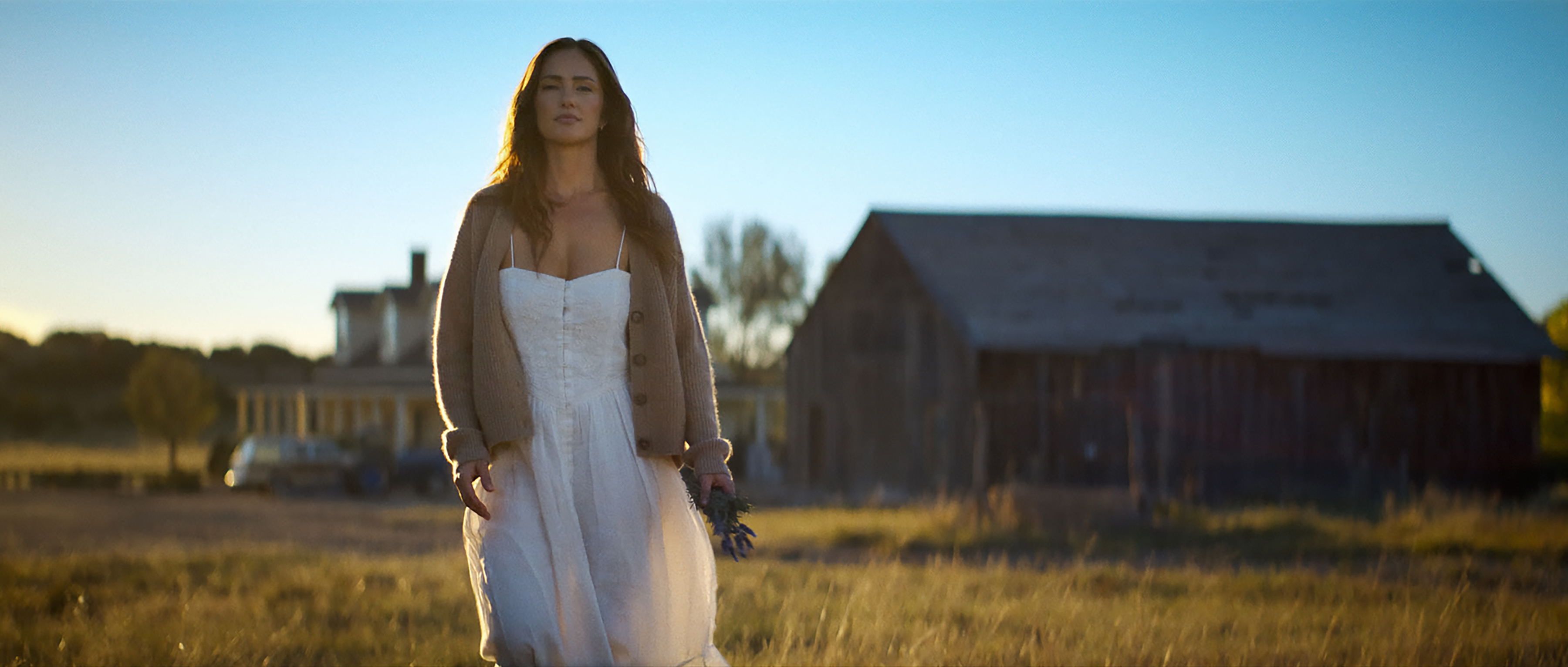 Minka Kelly as Quinn wearing a cardigan and white dress walking through a field by a bar in ransom canyon season 2