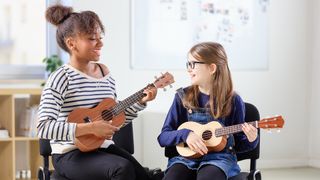 Two young girls sit side by side playing ukulele