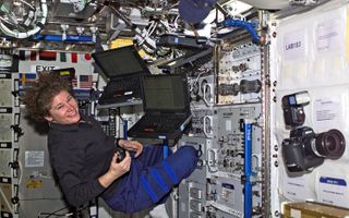 Susan Helms smiles and 'floats' in microgravity surrounded by two laptops and a camera.
