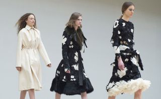 Three women on a catwalk, one in a cream coat, and two in black and white dresses