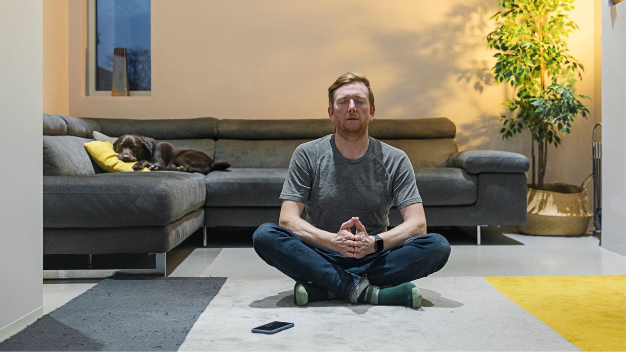 man wearing jeans and a tshirt in a living room setting in a crosslegged position meditating, facing the camera. there is a L-shaped sofa and plant behind him.