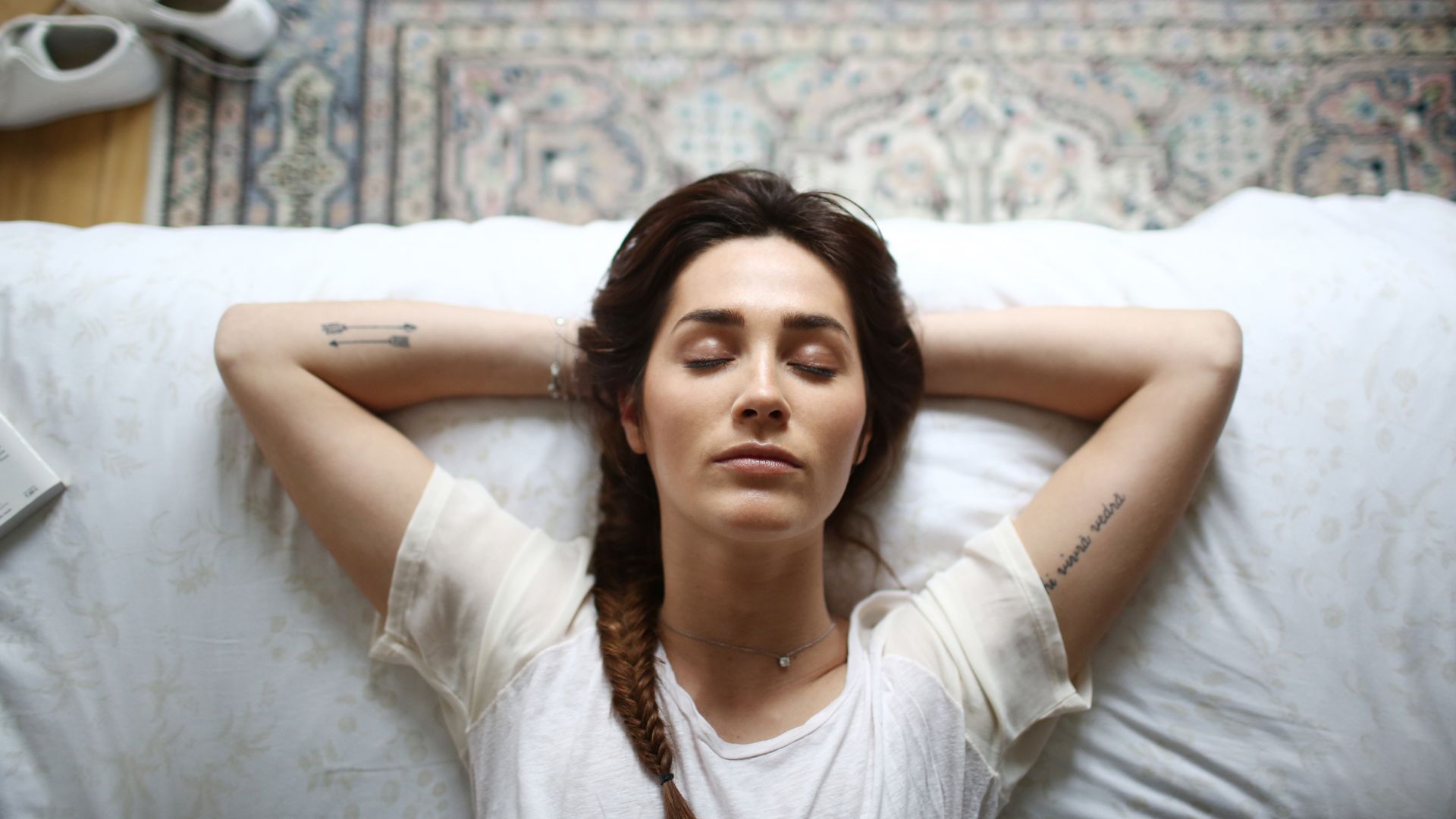 A woman lies on her back in bed with her arms behind her head. She has her eyes closed.