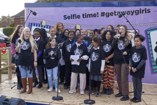The Getaway Girls Choir perform outside the new building on the big reveal day, surrounded by microphones and sound booms, standing in front of a purple banner that says "Selfie time! #getawaygirls"