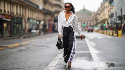 PARIS, FRANCE - JANUARY 23: Emilie Joseph wears sunglasses, a white oversized long shirt from & Other Stories, black bras from Aubade, a Danse Lente black leather bag, black and white striped Adidas flared jogger sport pants, fishnet tights from Calzedonia, black high heeled pointy shiny shoes from Cosmo Paris, on January 23, 2021 in Paris, France. (Photo by Edward Berthelot/Getty Images)