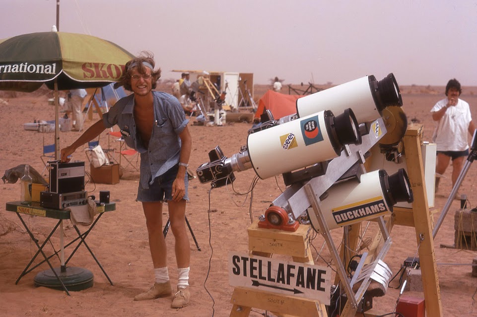a man smiles beside a telescope in the desert